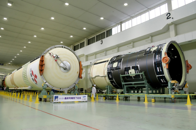 H2A最終50号機の第1段(左)と、第2段と黒い段間部が結合したもの。打ち上げ時、第1段は手前が上、第2段は奥が上となる。記念の横断幕が掲げられた=先月25日、愛知県飛島村の三菱重工業